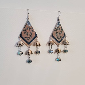 May include: A pair of dangle earrings with a diamond-shaped pendant featuring a floral pattern in shades of brown, green, and blue. The pendant is accented with gold-colored bells and blue crystal beads.
