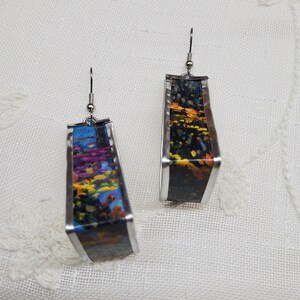 May include: A pair of silver earrings with a rectangular shape. The earrings are decorated with a colorful floral pattern on a black background.
