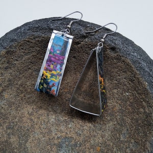 May include: A pair of earrings with a silver metal frame. One earring is rectangular and features a colorful abstract design. The other earring is triangular and has a black and white abstract design.