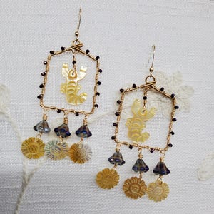 May include: A pair of gold-toned earrings with a rectangular frame design. Each earring features a yellow lobster charm, dark purple beads, and dangling flower and bell-shaped beads. The earrings are on a white background.