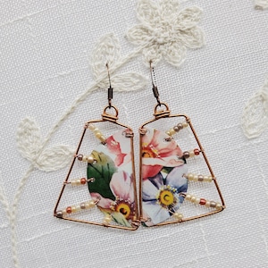 May include: A pair of copper wire earrings with a floral pattern and white beads. The earrings are shaped like a triangle with a rounded top.