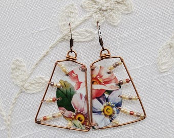 May include: A pair of copper wire earrings with a floral pattern and white beads. The earrings are shaped like a triangle with a rounded top.