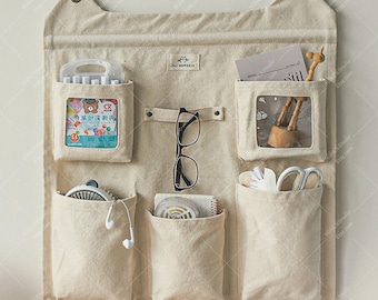 Puede incluir: Organizador de pared de tela beige con seis bolsillos de diferentes tamaños. El organizador tiene forma de orejas de gato en la parte superior e incluye un par de gafas, bolígrafos, tijeras y otros objetos pequeños. El organizador se fija a la pared con ventosas.