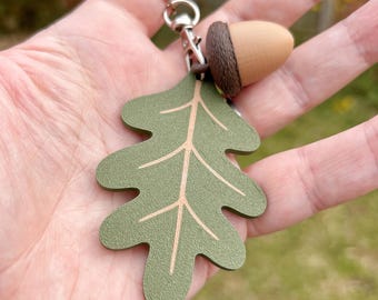 May include: A keychain featuring a green oak leaf and a wooden acorn charm. The leaf has light brown vein details, and the acorn has a brown cap. The keychain is attached to a silver-colored ring.