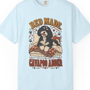 Puede incluir: Una camiseta azul claro con un gráfico de un perro Cavapoo blanco y negro acostado sobre una almohada con una manta a cuadros. El texto sobre el perro dice "BED MADE." y el texto debajo del perro dice "CAVAPOO ADDED." con pequeñas huellas.