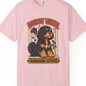 May include: A light pink short-sleeve t-shirt featuring a graphic of a black and tan dog sitting in a window frame. The text reads WINDOW WATCH above and NEIGHBORHOOD REVIEWED below. The design includes binoculars, paw prints, and a small bird.