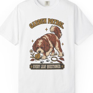 Puede incluir: Una camiseta blanca de manga corta con el gráfico de un perro marrón y blanco olfateando una hoja. El texto dice GARDEN PATROL y EVERY LEAF QUESTIONED. El diseño incluye una lupa, una flor y huellas de patas en tonos de color marrón.