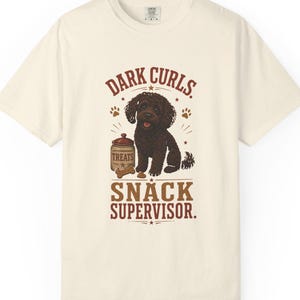 May include: A cream-coloured short-sleeve t-shirt featuring a graphic of a dark brown curly-haired dog sitting next to a jar labelled TREATS. The text reads DARK CURLS. SNACK SUPERVISOR. The design includes small paw prints and a bone illustration.