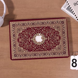 Può includere: Una skin per laptop con uno sfondo rosso intenso e un intricato motivo floreale dorato e nero che ricorda il design di un tappeto tradizionale. Il centro presenta un ritaglio circolare che rivela il logo Apple bianco sul coperchio.