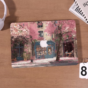 Può includere: Una skin per laptop con una scena pittoresca di un grazioso negozio blu con alberi in fiore rosa. Il design include una bicicletta, un piccolo cartello e persone sedute ai tavoli. Il logo Apple è visibile.