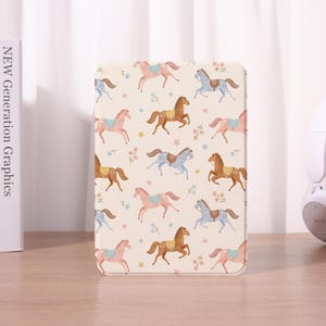 Może przedstawiać: Kremowe etui na tablet z powtarzającym się wzorem kreskówkowych koni w kolorze różowym, brązowym i niebieskim. Konie ozdobione są siodłami i otoczone małymi kwiatami i gwiazdami. Etui jest prostokątne z zaokrąglonymi rogami.