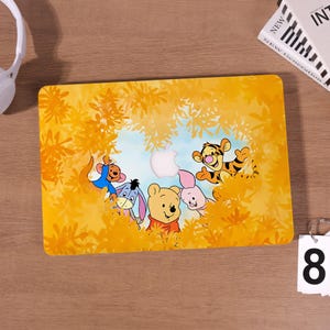 May include: A rectangular laptop hard shell case featuring a yellow-toned forest scene with Winnie the Pooh, Piglet, Tigger, Eeyore, and Roo. The design incorporates a central cutout for an Apple logo, surrounded by yellow foliage and characters.