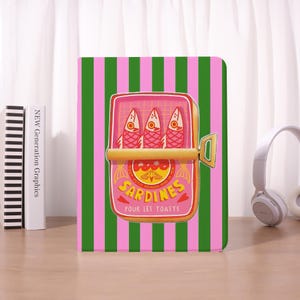 May include: A notebook with a pink and green striped background. The cover features a pink sardine can illustration with three fish and the text "Sardines Pour Les Toasts". A gold-colored pull tab is visible.