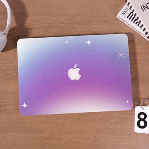 May include: A laptop skin featuring a colour gradient of blue, purple, and white. The Apple logo is in the centre. White starburst graphics are scattered across the surface. The skin is on a wooden surface.