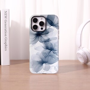 May include: A white phone case with a floral design in shades of blue and grey. The case has a glossy finish and a camera cutout. The phone case is on a wooden surface.