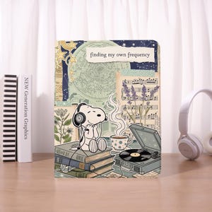Może przedstawiać: Dekoracyjny notatnik ze Snoopy'm w słuchawkach, z napisem "finding my own frequency". Projekt zawiera mapę w stylu vintage, nuty, lawendę i gramofon. Stos książek i filiżanka kawy dopełniają ilustrację.
