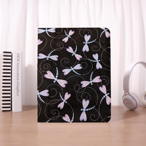 May include: A black notebook cover featuring a repeating pattern of lavender and light blue dragonflies with beaded accents. The dragonflies are set against a backdrop of swirling lines. The notebook is on a wooden surface.