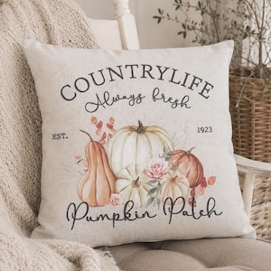 Könnte beinhalten: Ein quadratisches beiges Zierkissen mit einem Aufdruck von verschiedenen Kürbissen und Blumen. Der Text lautet "COUNTRYLIFE Always fresh" oben, "EST. 1923" in der Mitte und "Pumpkin Patch" unten in einer dunklen Schreibschrift.