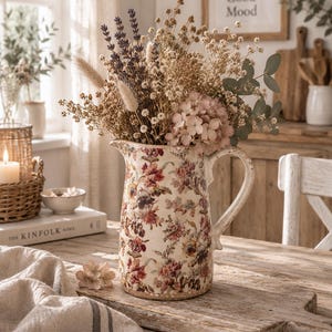 Könnte beinhalten: Ein Keramikkrug mit cremefarbenem Untergrund und einem mehrfarbigen Blumenmuster in Rot-, Braun- und Grüntönen. Der Krug ist mit einem Trockenblumenarrangement gefüllt, das Lavendel, Eukalyptus und hellrosa Hortensienblüten enthält.