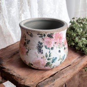 Könnte beinhalten: Runder, cremefarbener Keramik-Blumentopf mit Used-Look. Der Topf ist mit rosa Rosen und grünem Laub verziert. Der Rand ist hellbraun. Der Topf steht auf einer Holzoberfläche.
