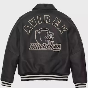 Puede incluir: Una chaqueta estilo universitario de color negro con un gran gráfico bordado en la espalda. El diseño incluye el texto AVIREX en arco sobre una cabeza de pantera negra rugiendo, con las palabras Black Aces en letra cursiva debajo.
