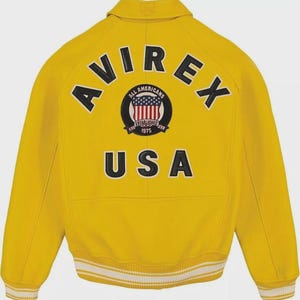 May include: A bright yellow varsity jacket featuring the text AVIREX in black arched lettering across the upper back. Below this is a circular patch with a shield graphic and the text ALL AMERICANS ESTABLISHED 1975. The word USA is in black.