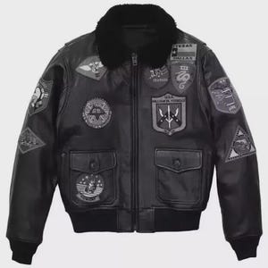 May include: A black leather bomber jacket featuring a black faux fur collar, a front zipper closure, and two flap pockets. The jacket is decorated with various embroidered patches, including a Texas flag, a U.S.S. William H. Standly shield, and a Tomcat emblem.