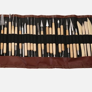May include: A brown leather tool roll containing a selection of sculpting tools. The tools feature wooden handles and metal tips, such as blades, loops, and modelling tools. The interior is black, and the roll has a snap closure.