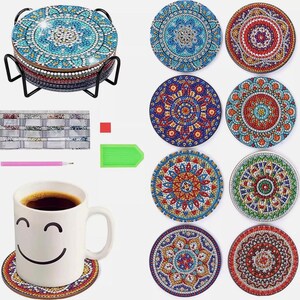 May include: A collection of mandala-patterned coasters, embellished with colourful, sparkling gems in blue, red, and gold. The coasters are displayed in a black metal stand. A white mug with a smiley face rests on one coaster.