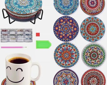 May include: A set of decorative coasters with a mandala design. The coasters are adorned with colorful, sparkling gems in shades of blue, red, and gold. A black metal stand holds the coasters. A white mug with a smiley face sits on one coaster.