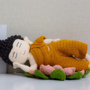 May include: A crocheted Buddha figure in a reclining pose, wearing a mustard-yellow robe. The figure rests on a pink and green lotus flower. The Buddha has black hair, closed eyes, and a small red smile. The background is a light neutral color.