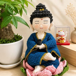 May include: A crocheted figurine of a seated figure with a black hair bun, wearing a dark blue robe with gold trim, resting on a pink and green crocheted lotus flower base. The figure has closed eyes, a small red dot on the forehead, and light skin.