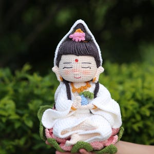 以下が含まれることがあります： 白いローブとピンクの花のついた頭飾りを身につけた、座っている姿の手作りのかぎ針編み人形。人形は黒髪で、額に赤い点があり、ピンクと緑の蓮の花の台座に座っています。