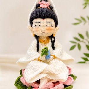 May include: A handcrafted crochet doll of a seated figure, wearing a white robe and a black head covering with a pink flower. The doll is seated on a lotus flower with pink and green petals, holding a small green and blue object.