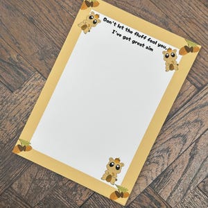 May include: A rectangular notepad with a yellow border and a white interior. The notepad features cartoon squirrels and acorns in each corner. Text on the notepad reads, "Don't let the fluff fool you, I've got great aim."