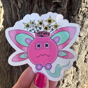 May include: A die-cut sticker featuring a round pink character with a wavy mouth, two purple circular feet, and a long light green tail. The character has large eyes with flower-shaped pupils and wings with pink and teal layers. Yellow flowers grow from the top.