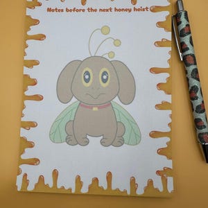 May include: A notepad with a cartoon bee-like creature and the text "Notes before the next honey heist." The notepad has a honey-dripping border. A pen with a leopard print design is next to the notepad.