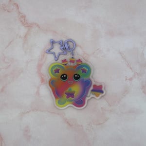 May include: A clear acrylic keychain featuring a tie-dye patterned bear character with large black eyes and a smiling mouth. The bear is decorated with small colorful stars and is attached to a light purple star-shaped metal lobster clasp.