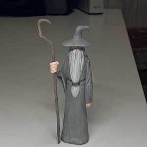 Puede incluir: Figura gris de un mago con una larga barba blanca, un sombrero puntiagudo y un bastón. El mago lleva una larga túnica gris y tiene las manos rosas. El bastón es marrón y curvado en la parte superior. La figura está sobre una superficie blanca.