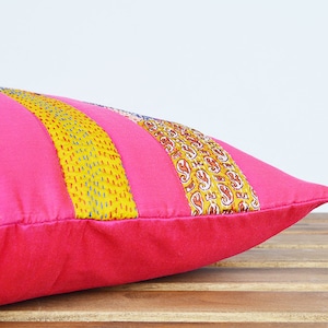 May include: A pink cushion with yellow, blue and brown patterned fabric strips. The cushion is on a wooden surface.