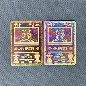 May include: Two rectangular trading cards featuring a holographic finish with a gold-toned outline of a creature resembling a cat. The cards display stylized symbols, geometric shapes, and non-standard text characters across the surface.