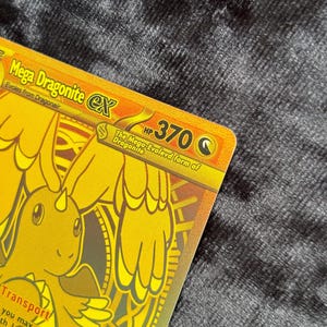 K&ouml;nnte beinhalten: Eine goldene Mega Dragonite EX Sammelkarte. Die Karte zeigt eine goldene Illustration eines drachenartigen Wesens mit dem Text "Mega Dragonite EX" und "HP 370". Die Karte enth&auml;lt auch den Text "Evolves from Dragonair".