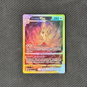 Op de afbeelding: Een holografische Arceus VSTAR Pokémon-kaart. De kaart heeft een gouden Arceus-ontwerp tegen een vurige rode en gele achtergrond. De kaart heeft de tekst "Trinity Nova" en "Starbirth".
