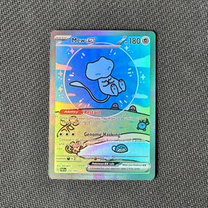 Op de afbeelding: Een holografische Mew ex ruilkaart met 180 HP. De kaart bevat een blauwe Mew-illustratie in een cirkelvormige bubbel, omringd door kleinere Pokémon-figuren op een regenboogkleurige achtergrond. De tekst bevat Ability Restart, Genome Hacking en Pokémon ex rule.