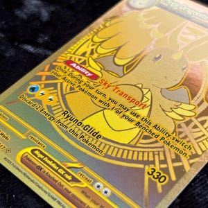 K&ouml;nnte beinhalten: Eine goldfarbene Mega-Dragonite-Pok&eacute;mon-Karte. Die Karte zeigt eine detaillierte Illustration eines drachenartigen Wesens mit dem Text "Sky Transport" und "Ryuno Glide". Die Karte enth&auml;lt auch die Nummer 330 und andere spielbezogene Texte.