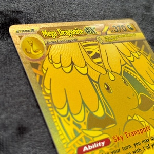 K&ouml;nnte beinhalten: Eine goldfarbene Mega Dragonite EX Sammelkarte. Die Karte zeigt eine detaillierte Illustration eines drachenartigen Wesens mit dem Text "Mega Dragonite EX" und "Sky Transport". Die Karte hat eine Stufe 2-Bezeichnung und 370 HP.