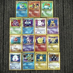 May include: A collection of fifteen Pokémon trading cards, featuring various characters with colorful illustrations and text. The cards are arranged on a gray surface, showcasing a variety of card designs and HP values.