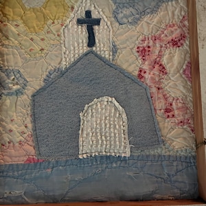 May include: A textile wall hanging featuring a denim church appliqué with a cross on the steeple and a textured white arched doorway. The church is centered on a background of vintage-style hexagonal patchwork quilt pieces in yellow, blue, and pink.