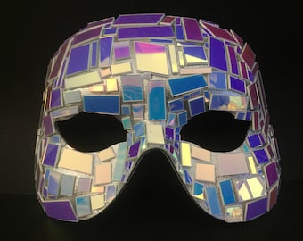 May include: A decorative mask with a mosaic design. The mask is covered in small, irregularly shaped iridescent tiles in shades of purple, blue, and white. The eye openings are dark, and the mask is set against a black background.