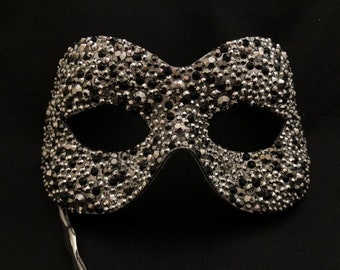 Può includere: Una maschera da ballo argentata con strass neri che coprono l'intera superficie. La maschera ha un'unica apertura per l'occhio e un lungo nastro sottile argentato.